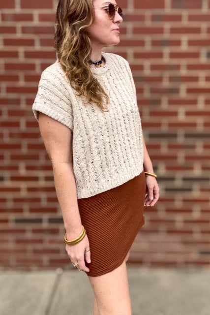 Woman wearing a beige knitted top and brown skirt against a brick wall.