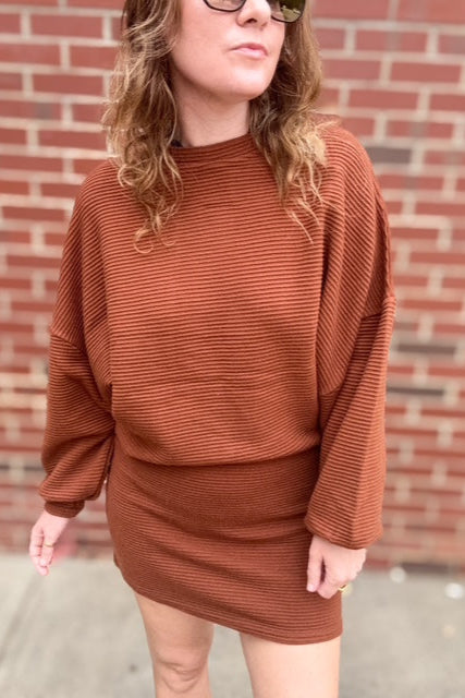 Person wearing a brown ribbed dress against a brick wall.
