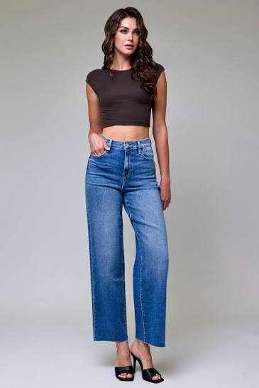 A woman modeling a pair of high-waisted, wide-leg blue jeans with a dark brown cropped top and black heels, standing against a neutral gray studio background.