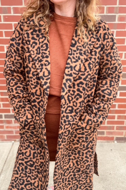 Person wearing a leopard print coat over a brown outfit against a brick wall.
