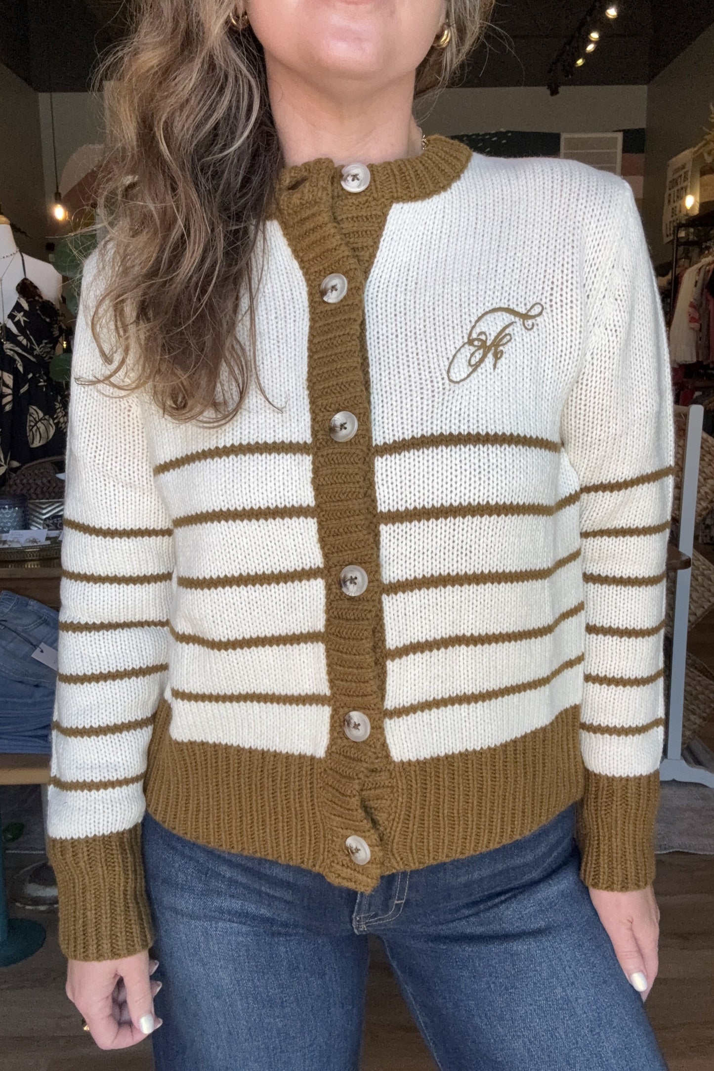 A woman wearing a cream and brown striped knit cardigan with a brown button placket and a brown embroidered logo on the chest, paired with blue jeans in a boutique setting.