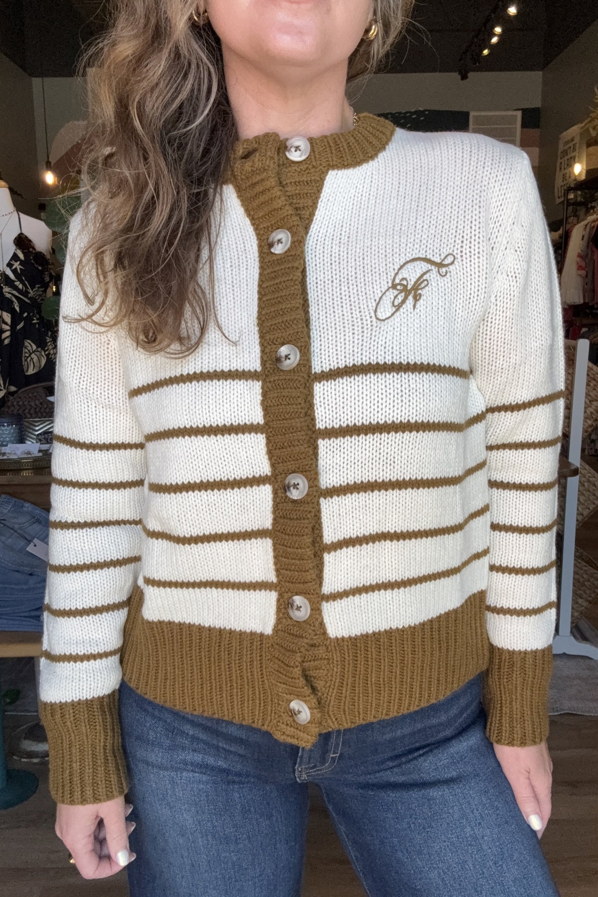 A woman wearing a cream and brown striped knit cardigan with a brown button placket and a brown embroidered logo on the chest, paired with blue jeans in a boutique setting.