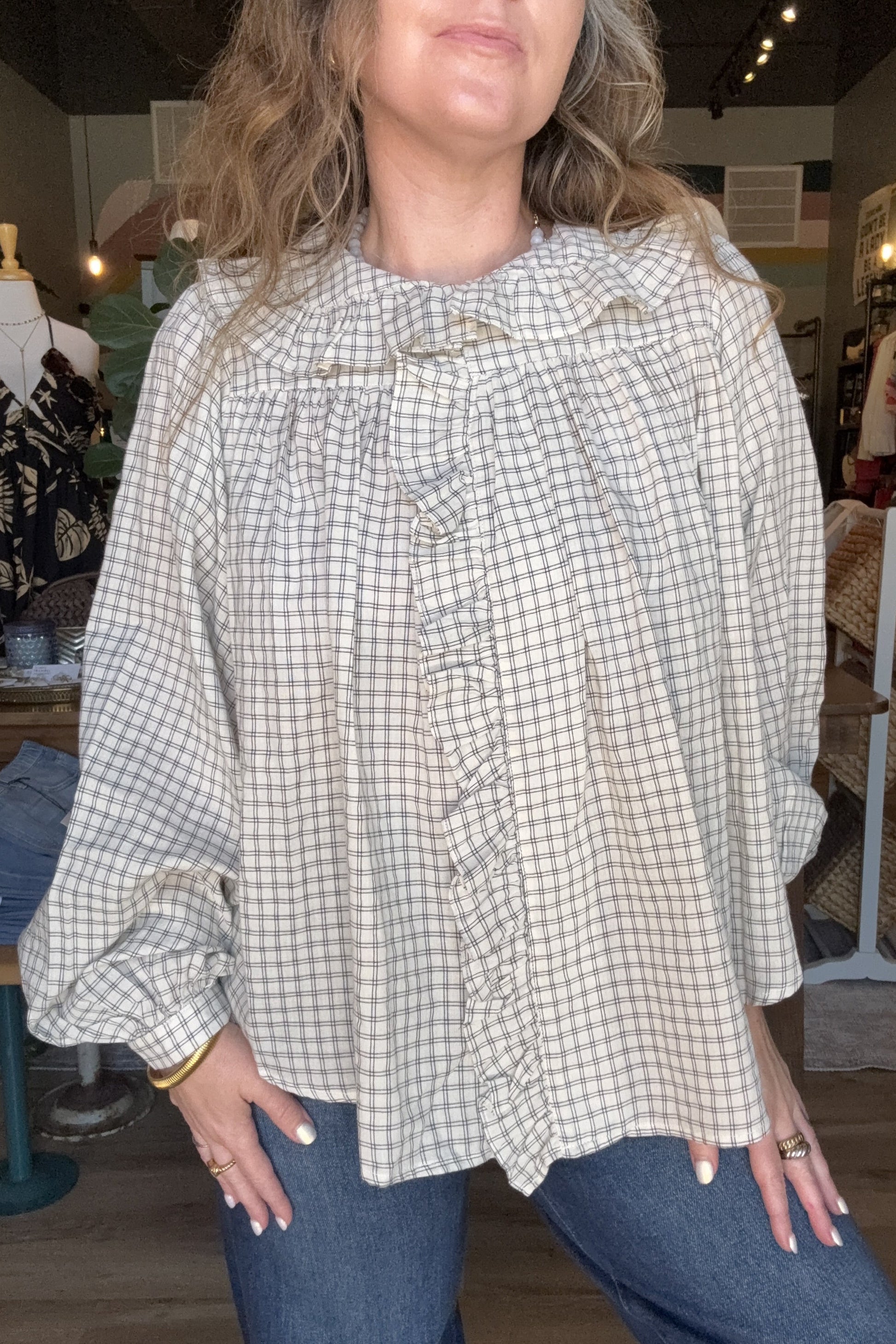 A woman wearing a beige and white gingham long-sleeve blouse with a ruffled front and gathered shoulders, paired with blue jeans in a clothing store setting.