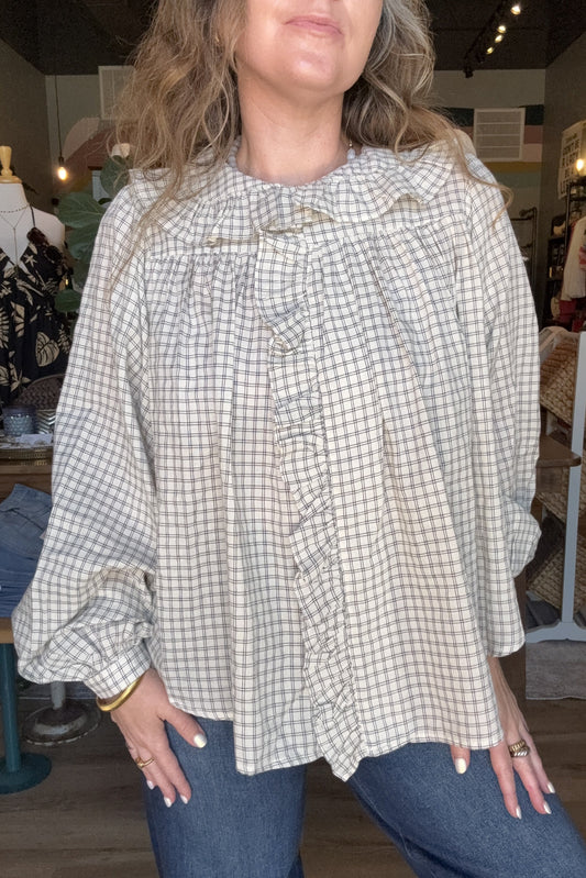 A woman wearing a beige and white gingham long-sleeve blouse with a ruffled front and gathered shoulders, paired with blue jeans in a clothing store setting.