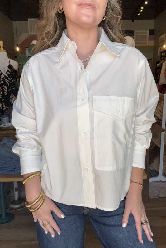 A woman wearing a white long-sleeve button-up shirt with a chest pocket, paired with blue jeans, standing inside a clothing boutique.