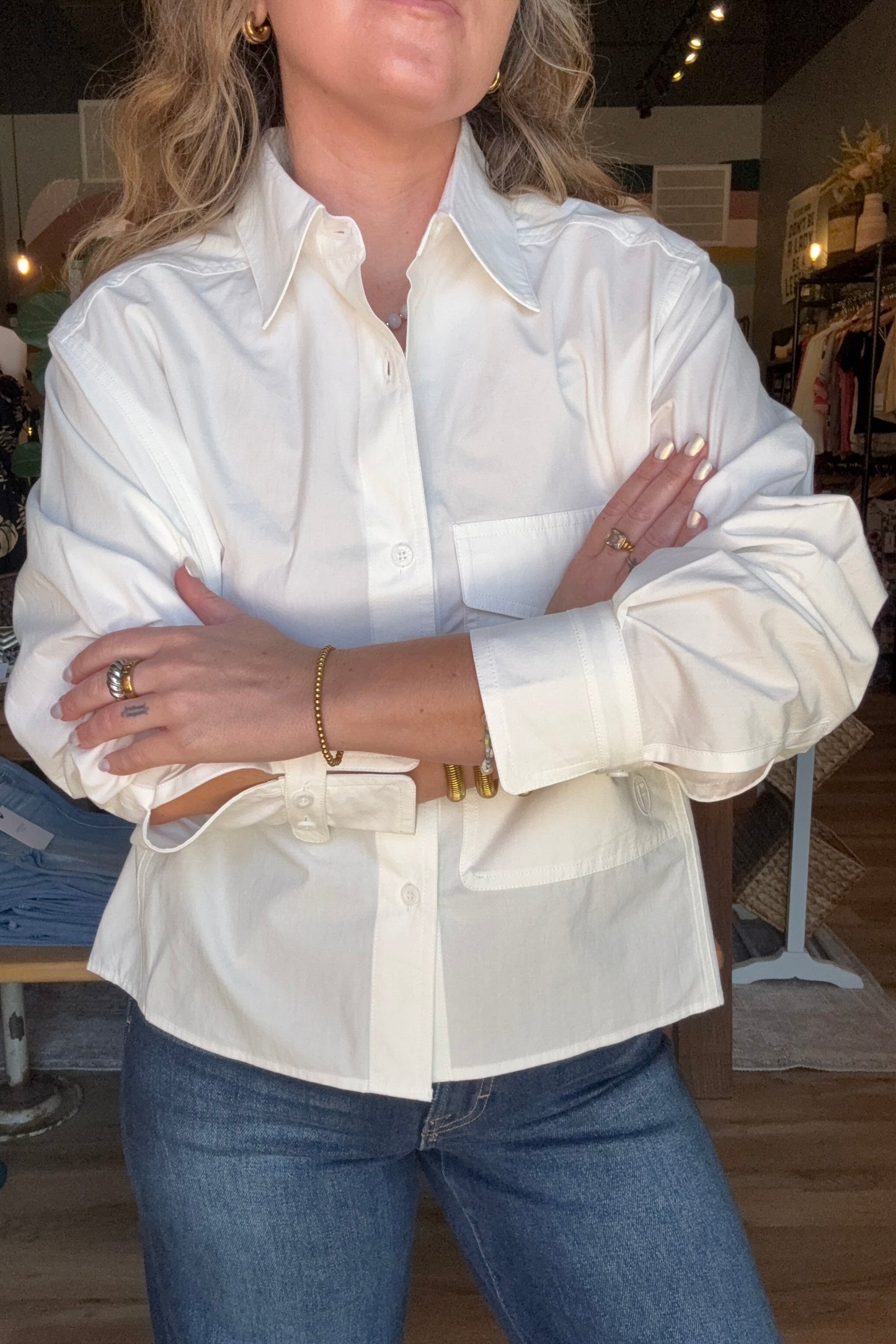 A woman wearing a white long-sleeve button-up shirt with a chest pocket and a matching belt, paired with blue jeans, standing in a clothing store.