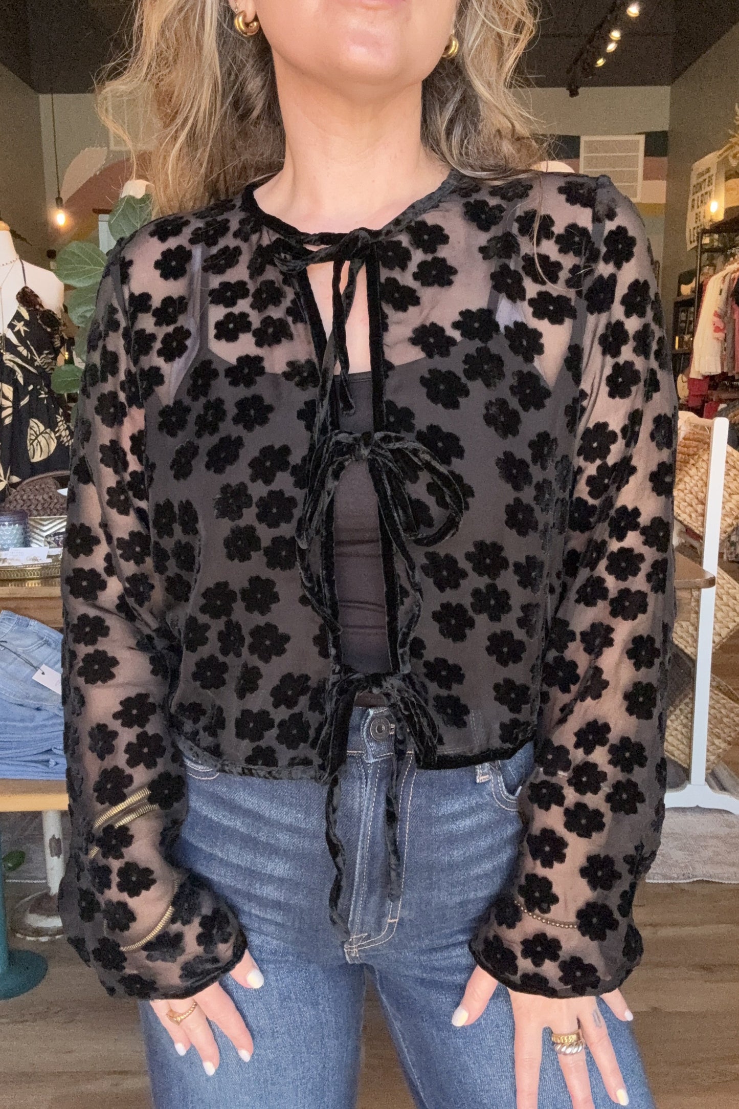 A woman wearing a sheer black long-sleeve top with a black floral pattern over a dark gray top, paired with blue jeans, in a clothing store.