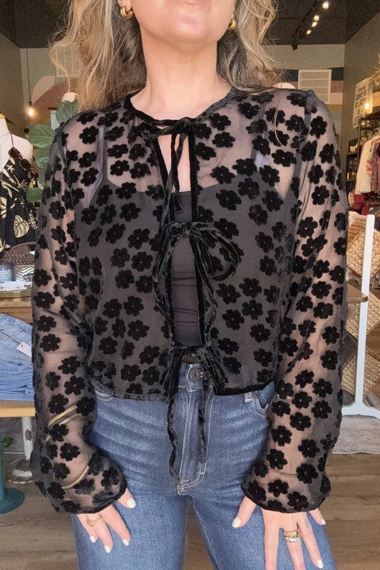 A woman wearing a sheer black long-sleeve top with a black floral pattern over a dark gray top, paired with blue jeans, in a clothing store.