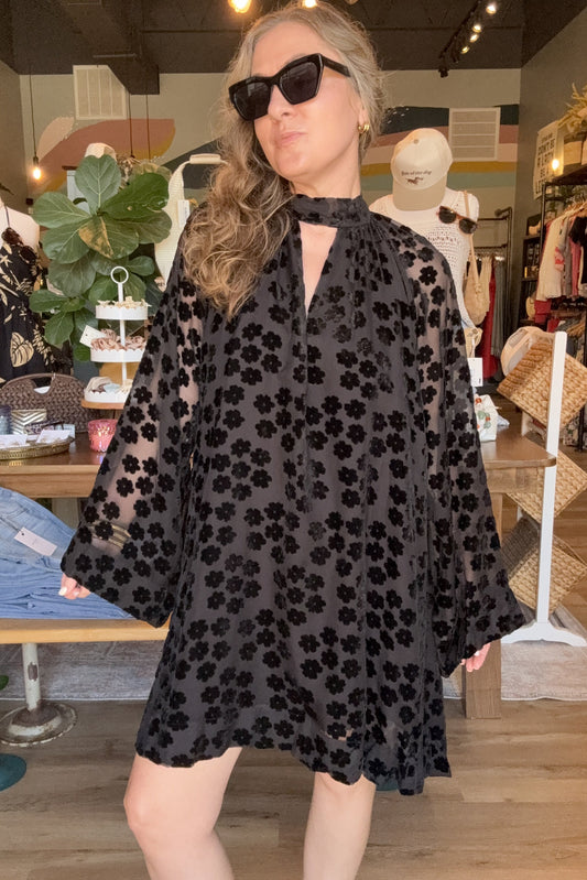 A woman wearing a sheer black floral mini dress with wide bell sleeves and a keyhole neckline, standing in a clothing boutique.