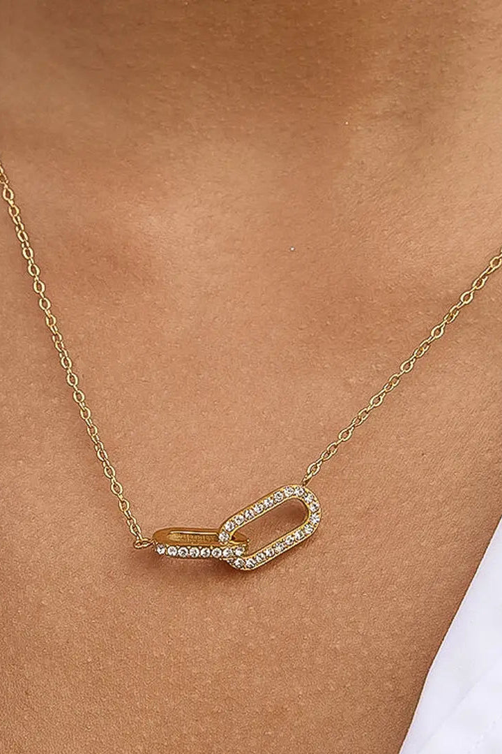 A gold chain necklace with a double-link pendant, one of which is paved with small clear stones, worn on a model's neck over a white top.