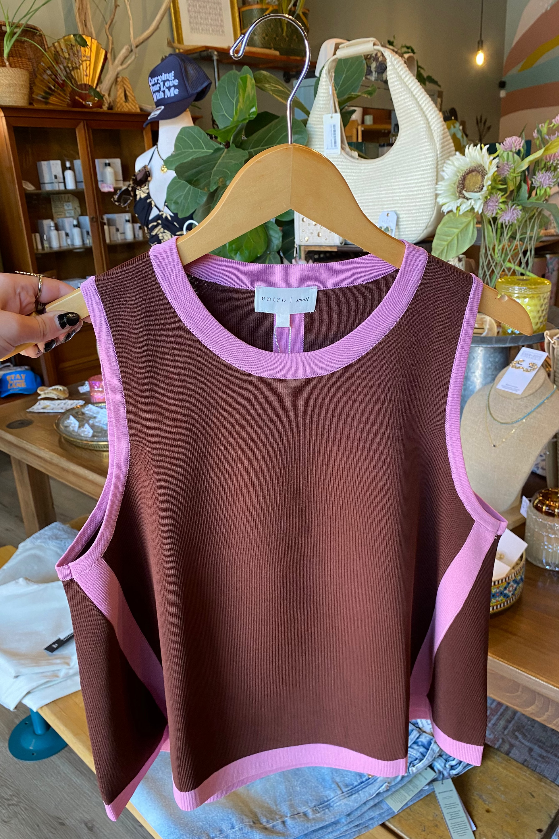 A sleeveless knit top in dark brown with light pink trim, hanging on a wooden hanger in a boutique. A hand is holding the left side of the garment.