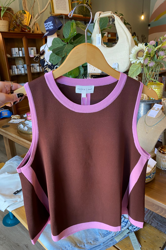 A sleeveless knit top in dark brown with light pink trim, hanging on a wooden hanger in a boutique. A hand is holding the left side of the garment.