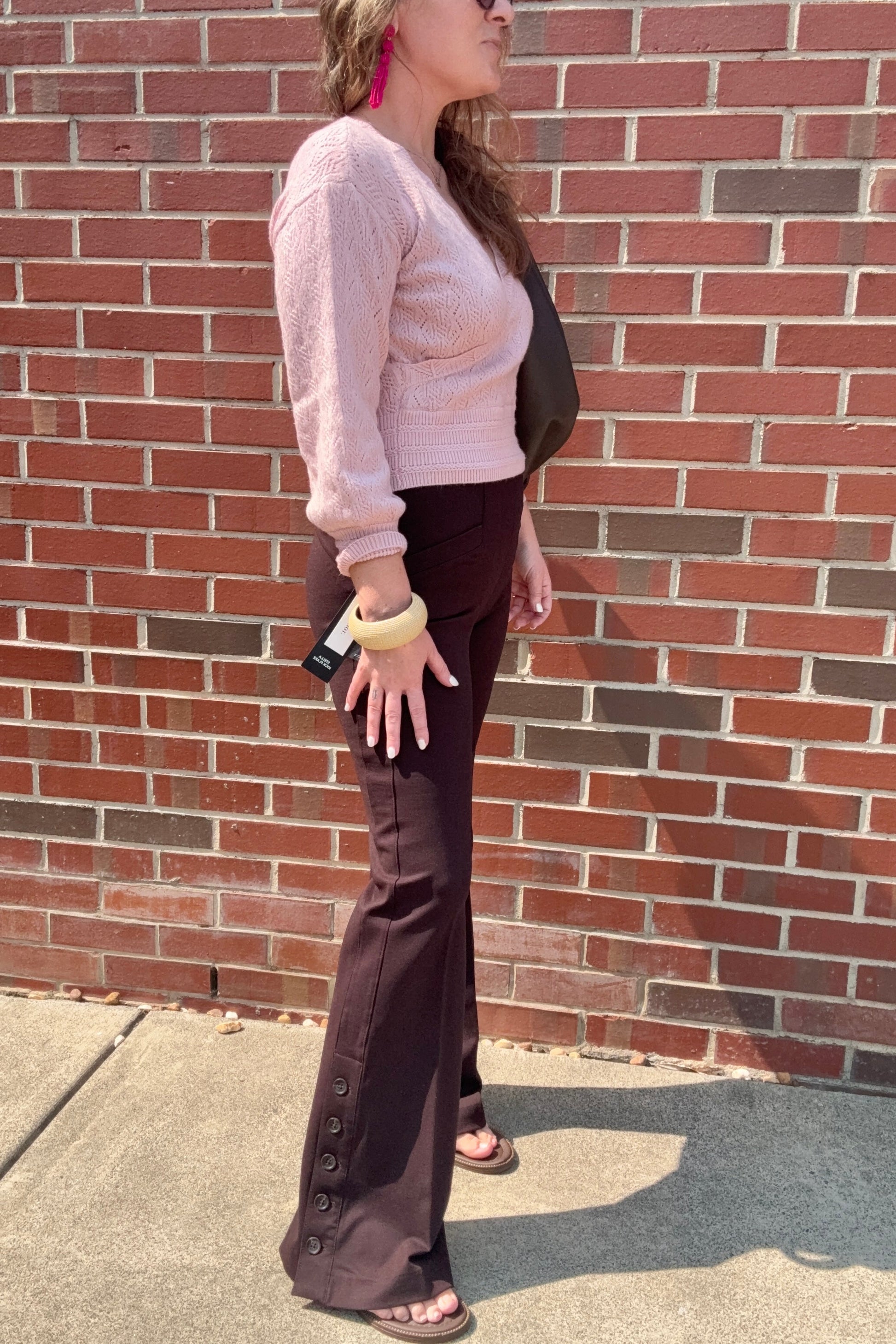 A woman stands in profile against a red brick wall, wearing a light pink fuzzy long-sleeve top, dark brown flared pants with decorative buttons at the hem, and brown sandals.