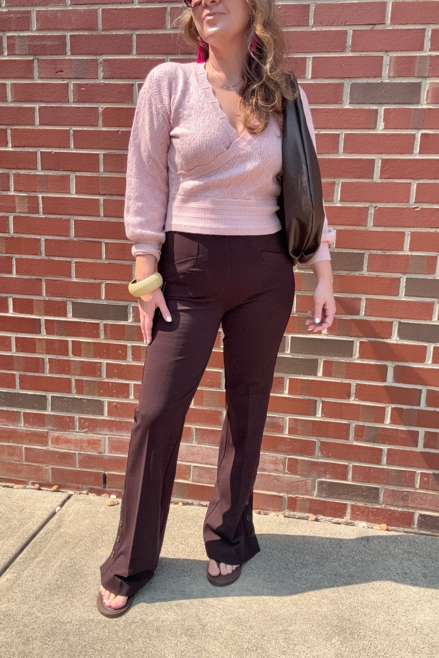 A woman stands outdoors against a red brick wall wearing a light pink knit wrap top, dark brown flared pants, and brown sandals, with a black shoulder bag.