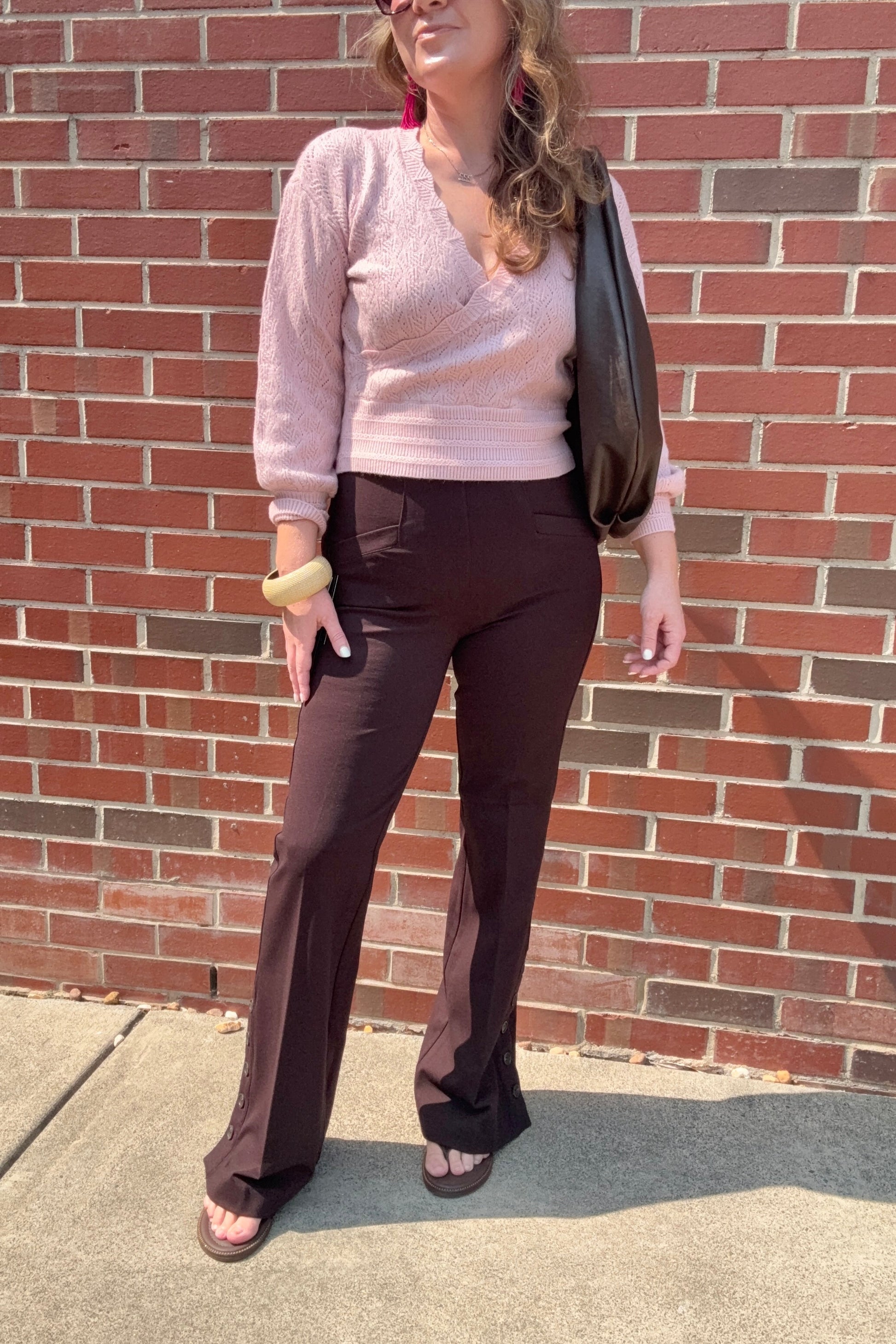 A woman stands outdoors against a red brick wall wearing a light pink knit wrap top, dark brown flared pants, and brown sandals, with a black shoulder bag.