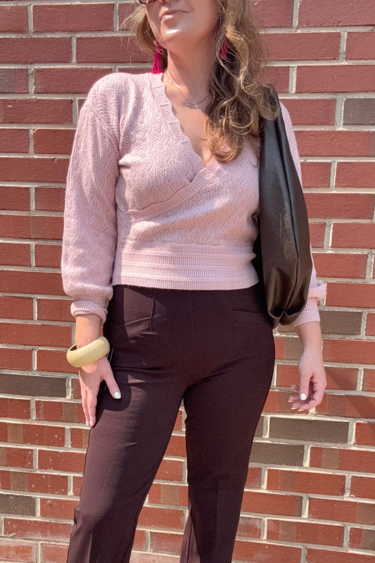 A woman wearing a light pink ribbed knit wrap sweater and dark brown pants, standing against a red brick wall and carrying a black shoulder bag.