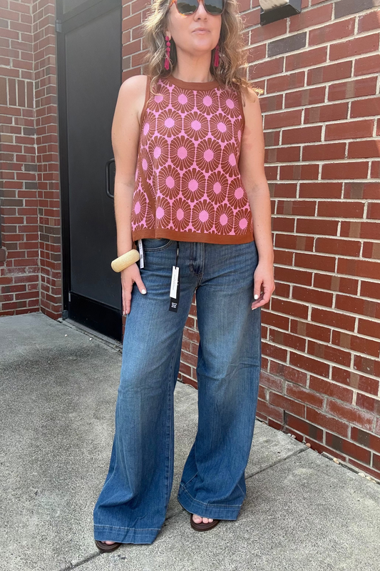 A woman wearing a sleeveless rust-brown knit top with a pink geometric pattern and wide-leg blue jeans, standing outdoors against a red brick wall.