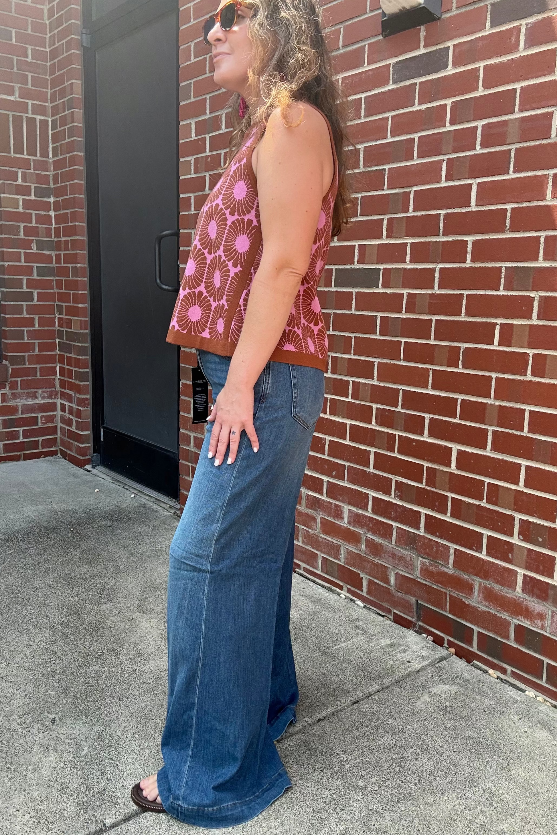 A woman stands in profile outdoors against a red brick wall, wearing a sleeveless pink patterned top and wide-leg blue jeans.