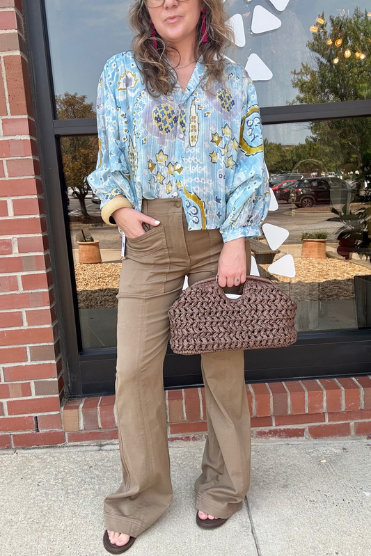 A woman stands outdoors in front of a brick storefront window wearing a light blue patterned blouse, tan wide-leg pants, and holding a brown woven handbag.