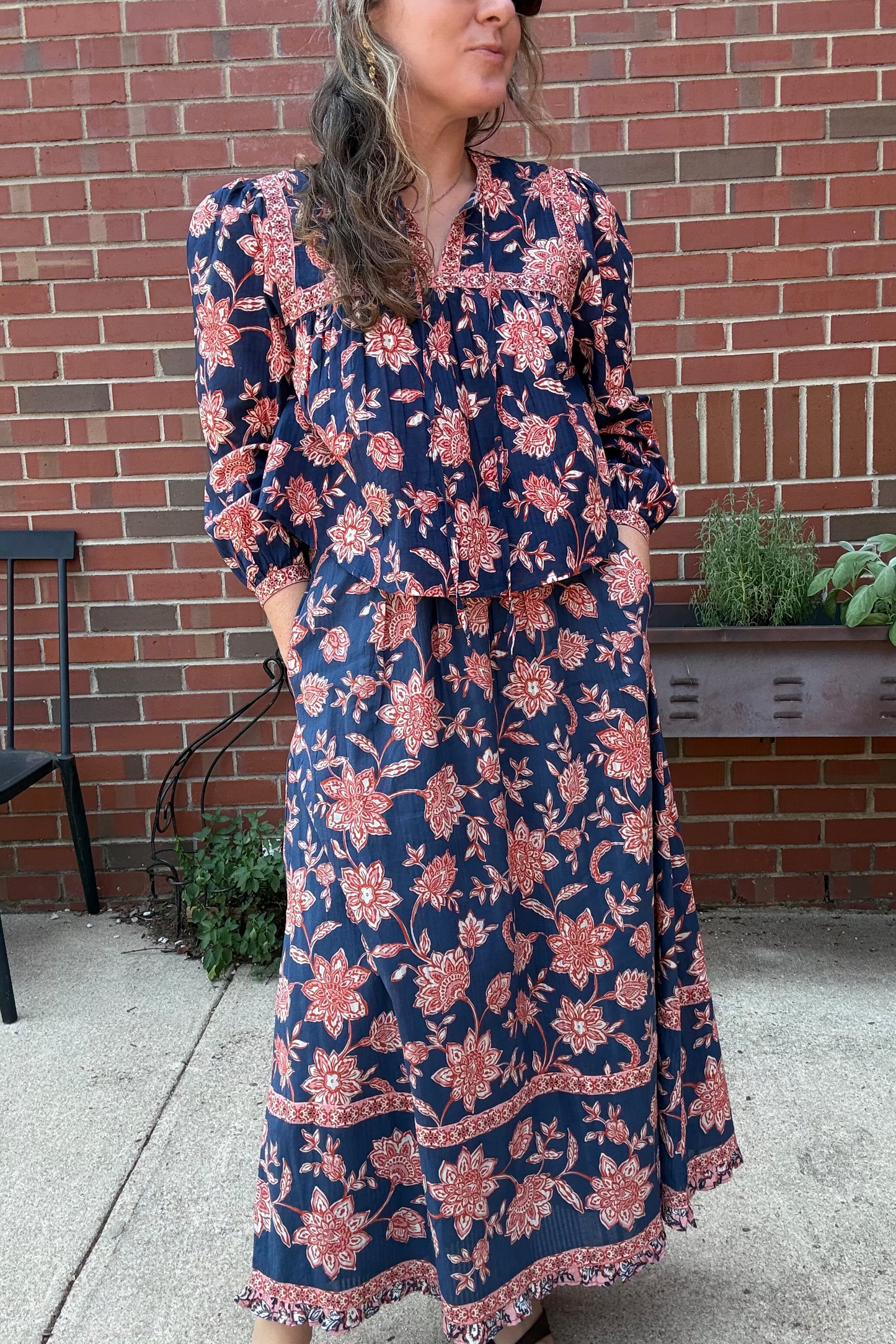 A woman wearing a navy blue maxi dress with a pink and orange floral print, standing outdoors in front of a red brick wall.