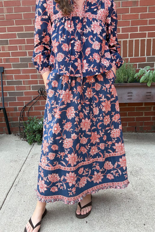 A woman wearing a navy blue maxi dress with a pink and coral floral print, standing outdoors on a concrete patio in front of a red brick wall.