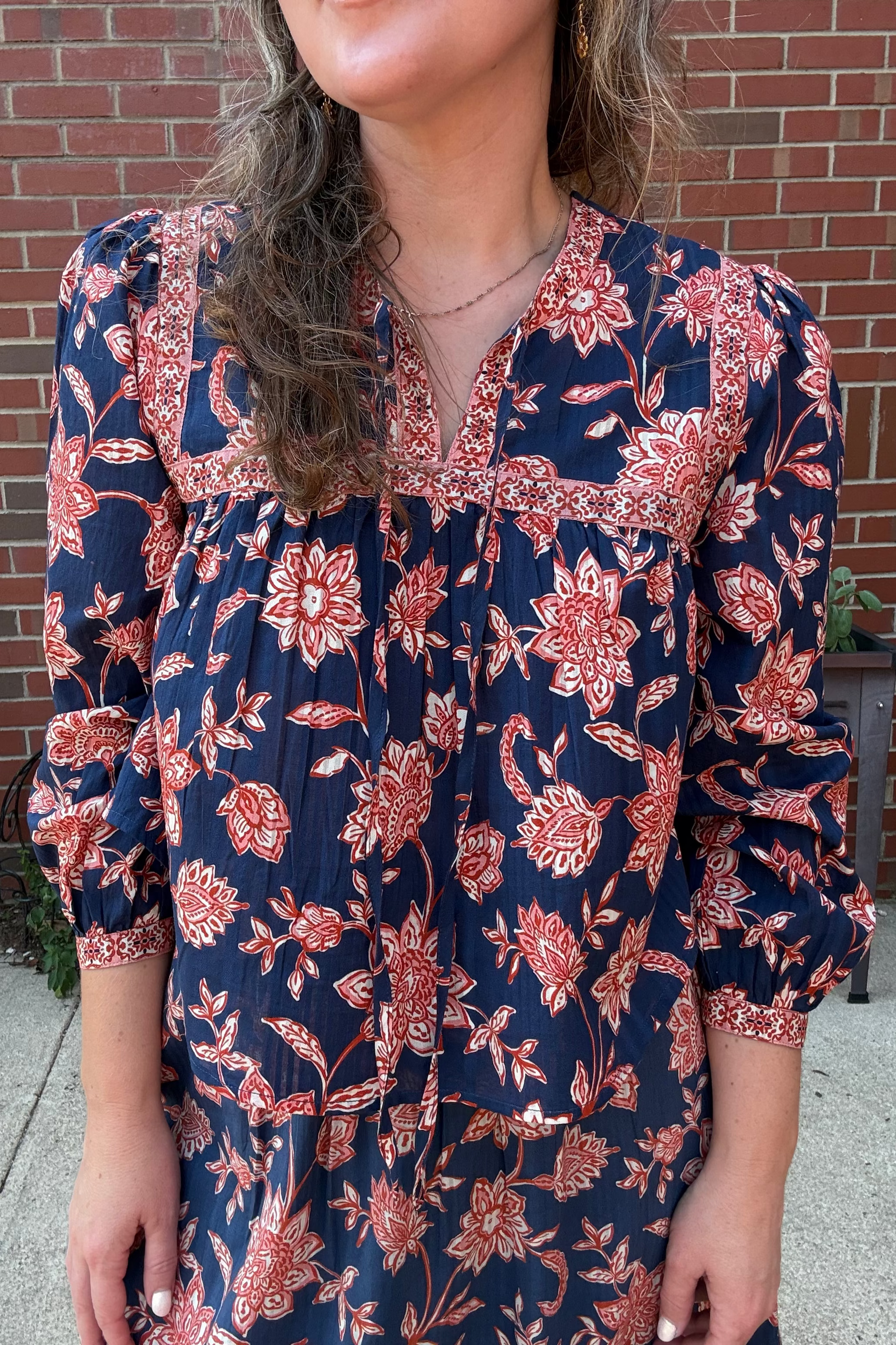 A woman wearing a navy blue long-sleeve blouse with a red and orange floral print, standing in front of a red brick wall.