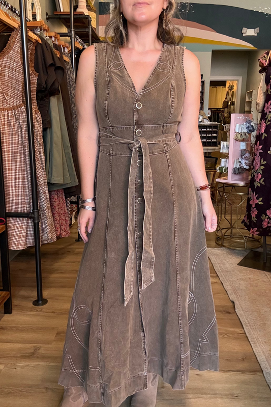 A woman wearing a sleeveless, V-neck, button-front maxi dress in a brownish-gray washed fabric with a matching tie belt, standing inside a clothing boutique.