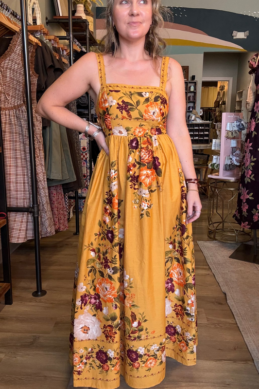 A woman wearing a mustard yellow floral maxi dress with a smocked bodice and wide straps, standing inside a clothing boutique.