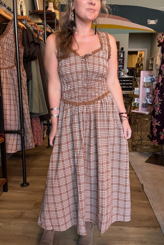 A woman modeling a sleeveless brown and beige plaid midi dress with lace-up front and ruffle trim inside a clothing boutique.