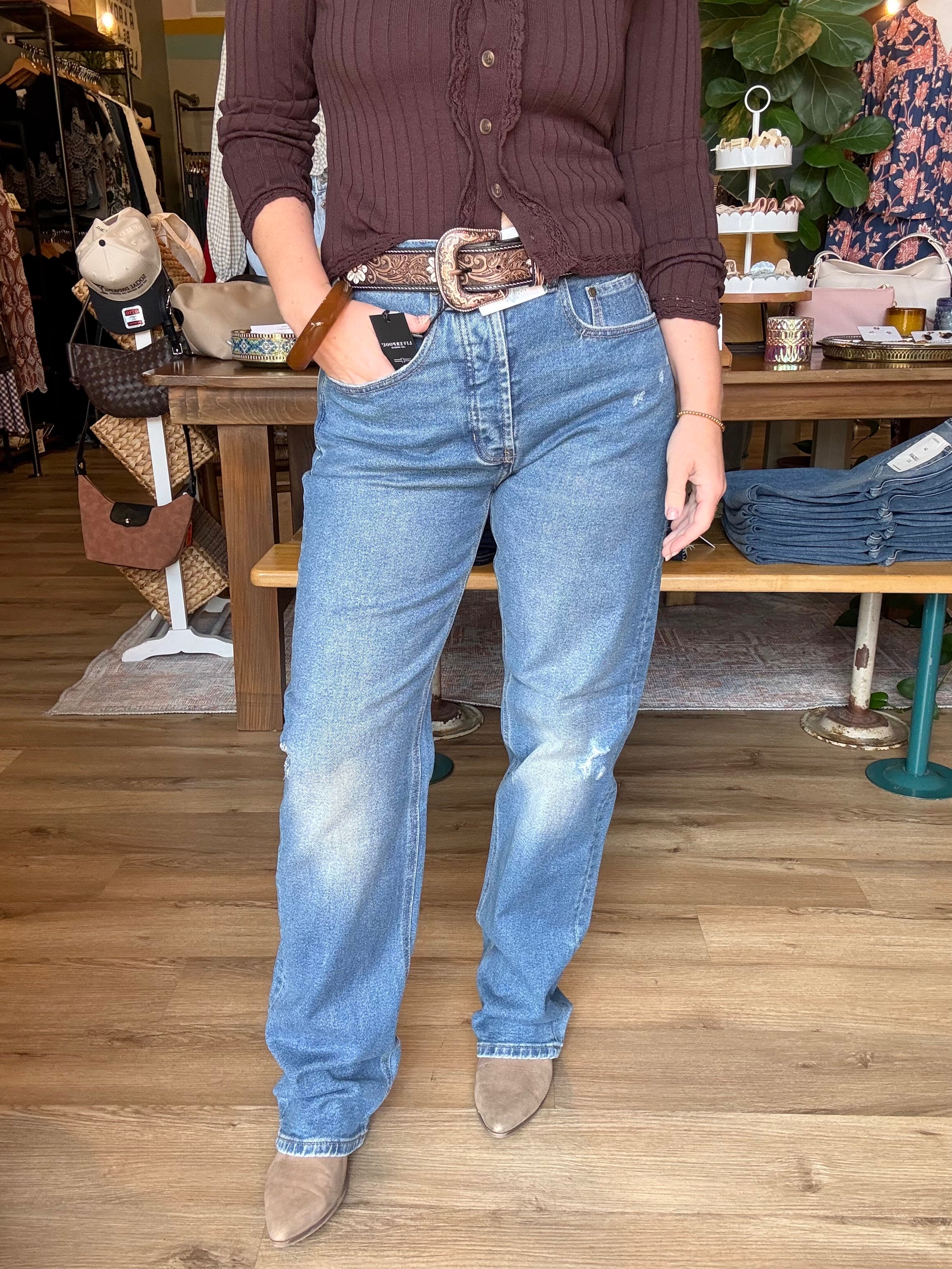 A person standing in a clothing store wearing light-wash blue jeans with a brown leather belt, a dark brown ribbed button-up top, and taupe pointed-toe boots.