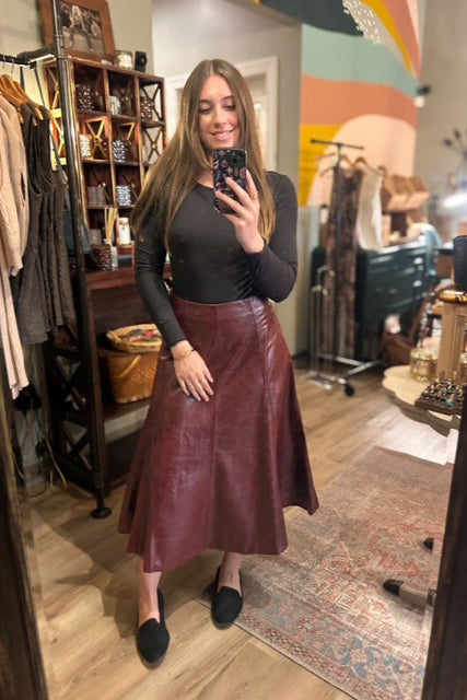 Person taking a mirror selfie wearing a black top and burgundy leather skirt in a clothing store.