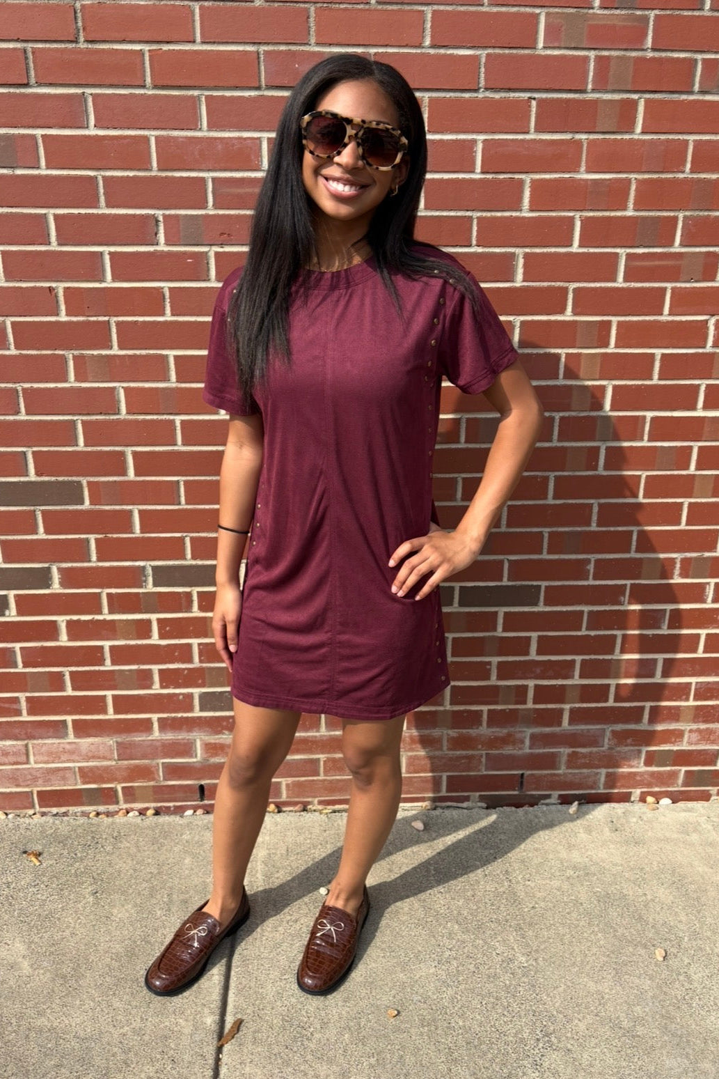 A woman wearing a short-sleeve burgundy t-shirt dress and brown leather loafers stands in front of a red brick wall.