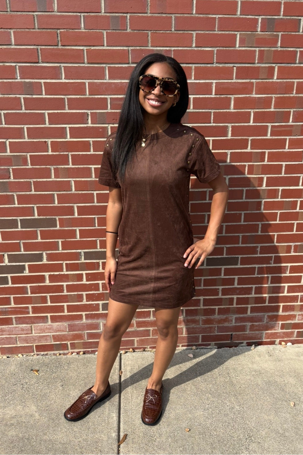 A woman wearing a brown short-sleeve dress and matching brown loafers stands in front of a red brick wall, wearing dark sunglasses.