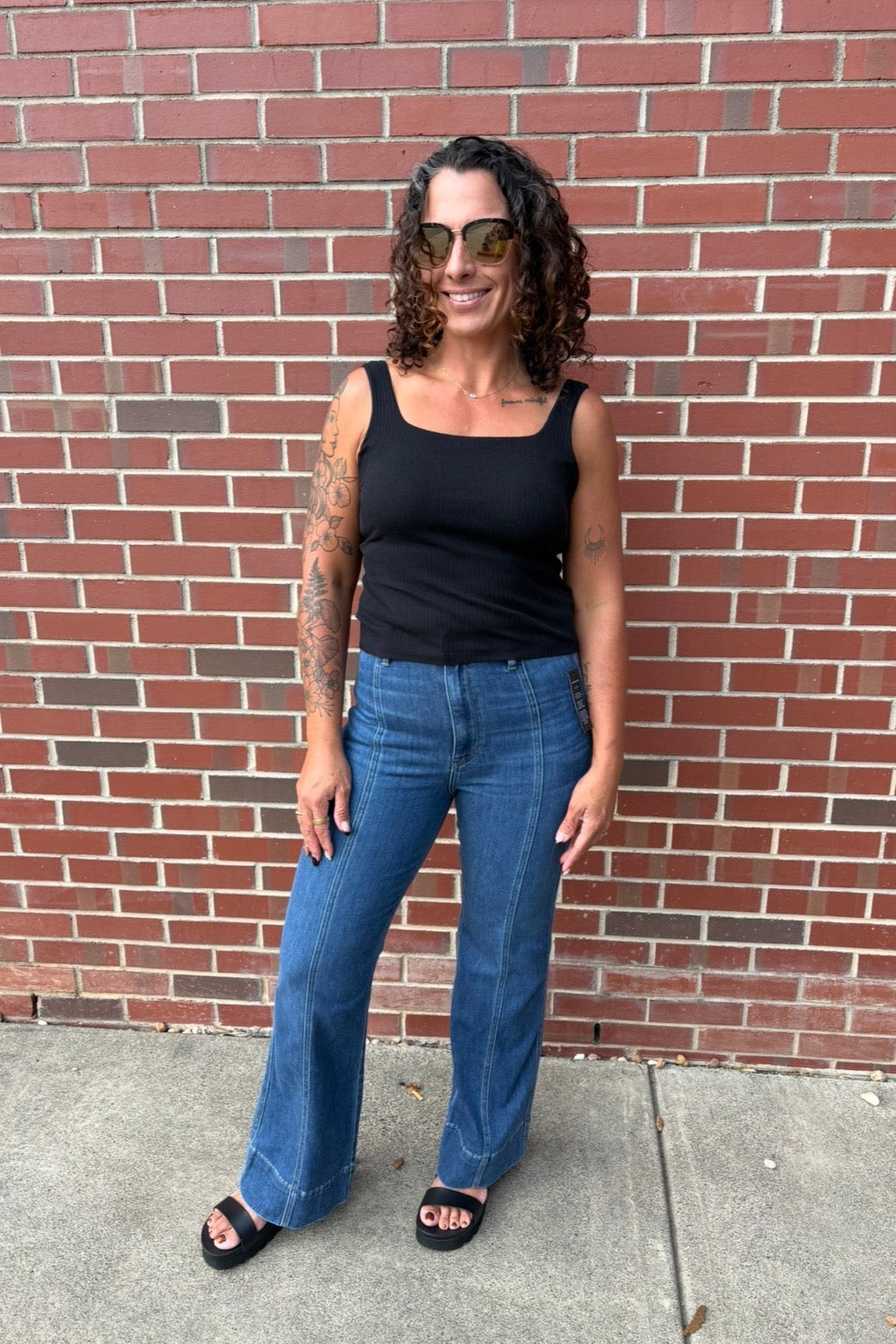 A woman with curly hair and sunglasses stands in front of a red brick wall, wearing a black tank top, blue jeans, and black slide sandals.