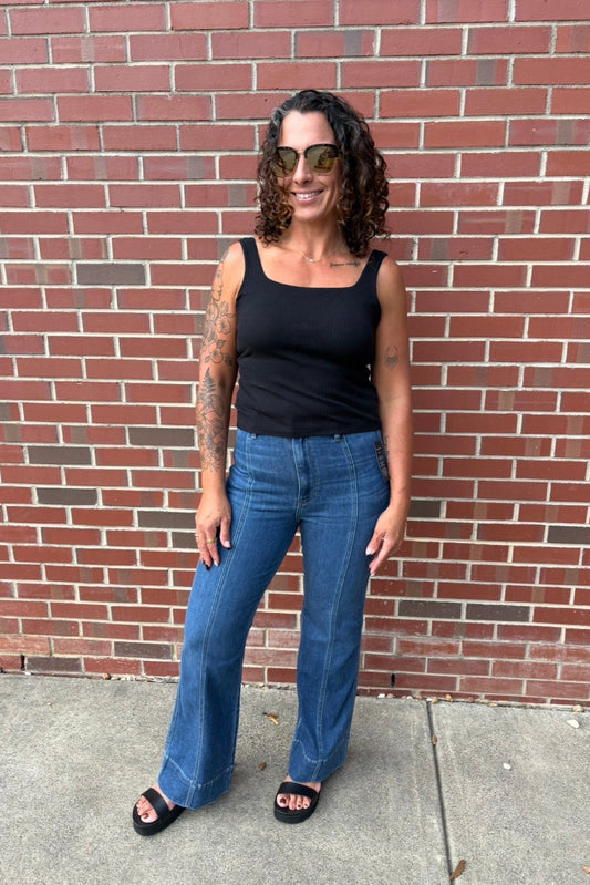 A woman with curly hair and sunglasses stands in front of a red brick wall, wearing a black tank top, blue jeans, and black slide sandals.