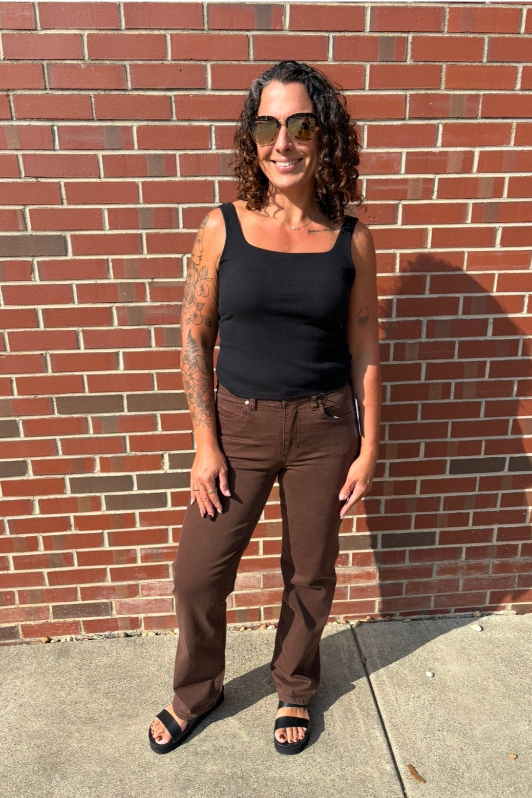 A woman with curly hair and sunglasses stands outdoors against a red brick wall, wearing a black tank top, brown pants, and black sandals.