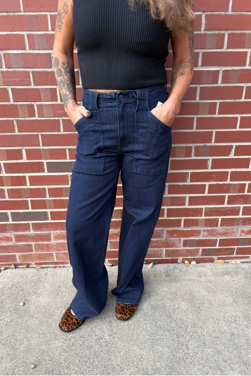 A person wearing dark blue wide-leg denim jeans with a black ribbed top and leopard print shoes, standing in front of a red brick wall.
