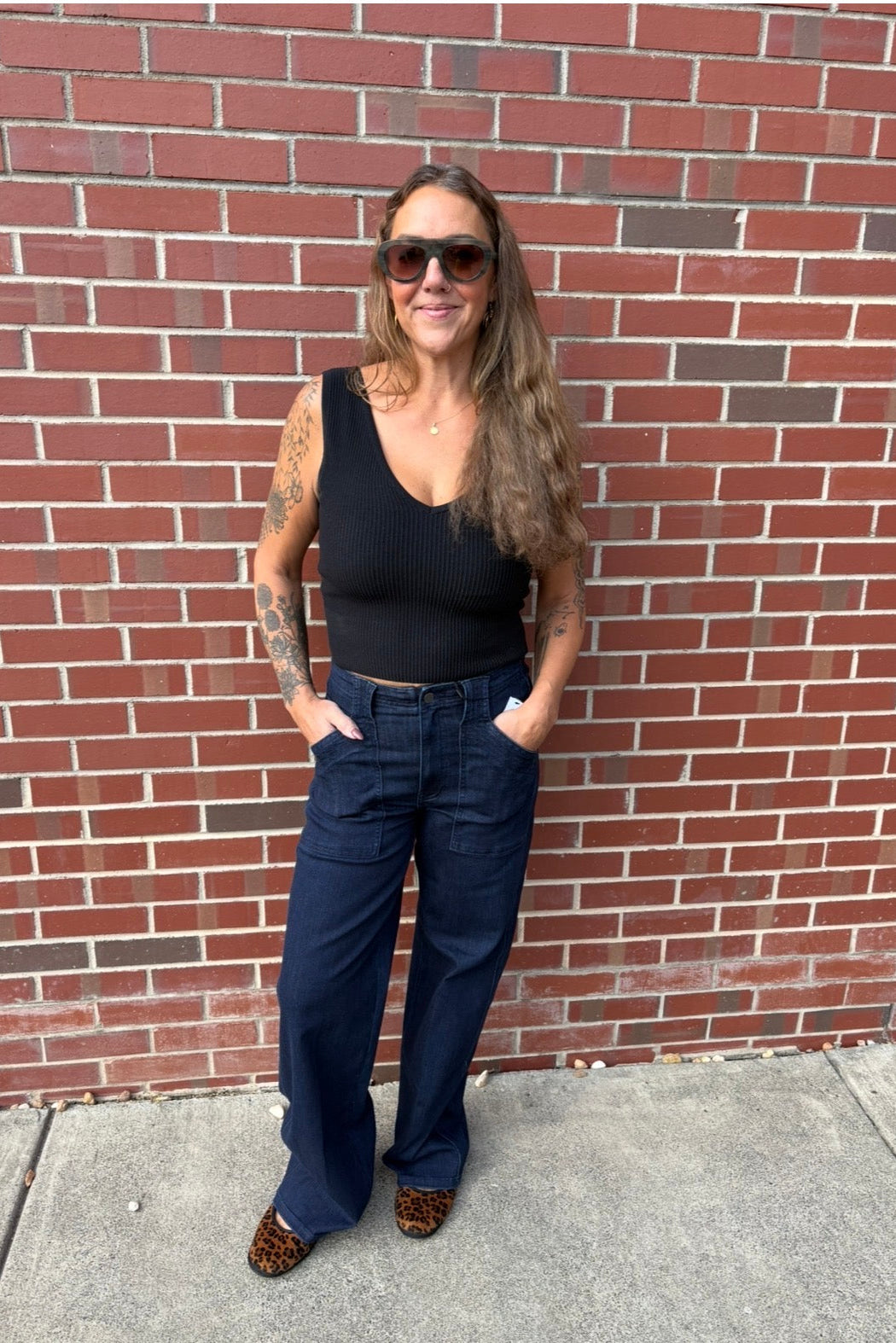 A woman stands against a red brick wall wearing a black ribbed tank top, dark blue wide-leg jeans, and leopard print shoes.