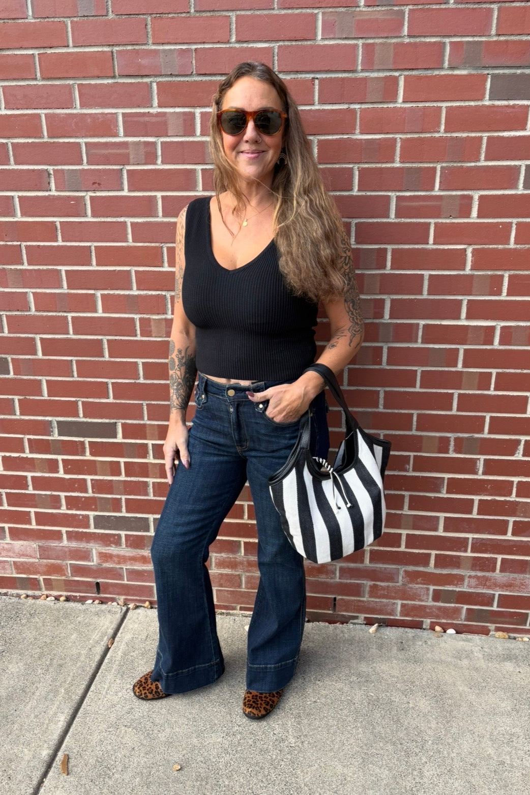 A woman stands against a red brick wall wearing a black ribbed tank top, dark blue flare jeans, leopard-print shoes, and sunglasses, while carrying a black and white striped tote bag.