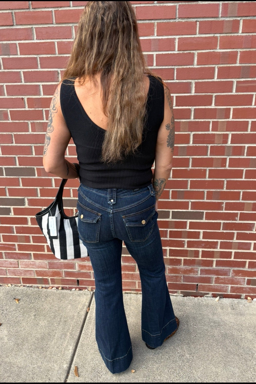 A woman viewed from behind stands on a sidewalk in front of a red brick wall, wearing a black ribbed tank top and dark blue flared jeans, with a black-and-white striped tote bag.