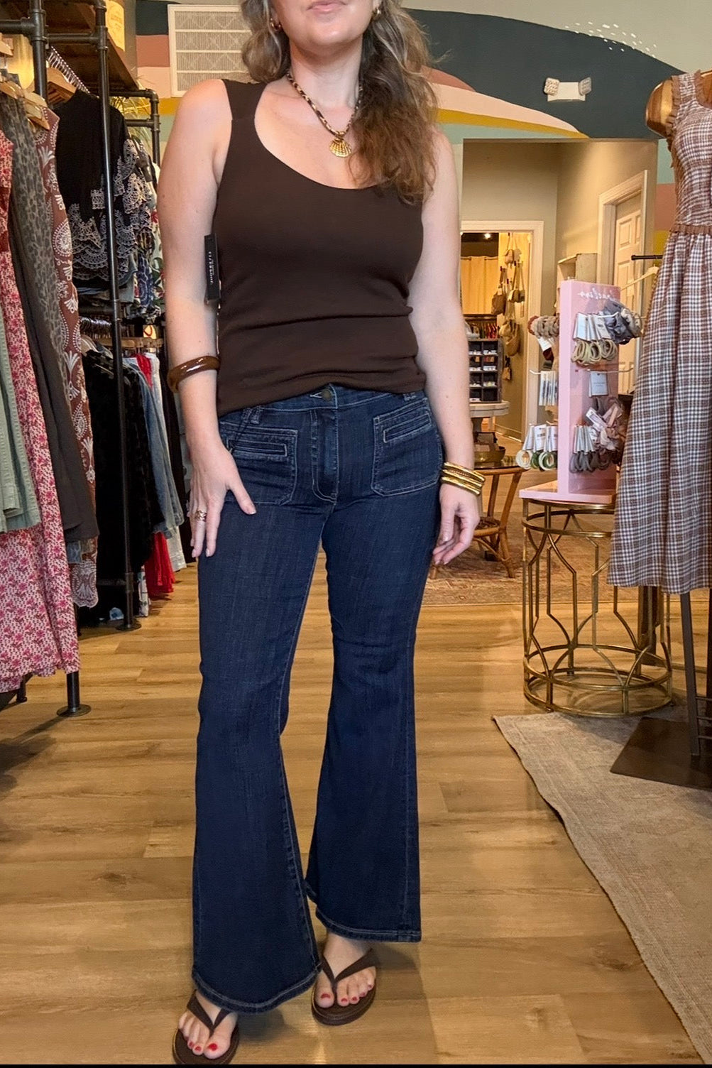 A woman modeling dark blue flare jeans with a brown scoop-neck tank top inside a clothing boutique.