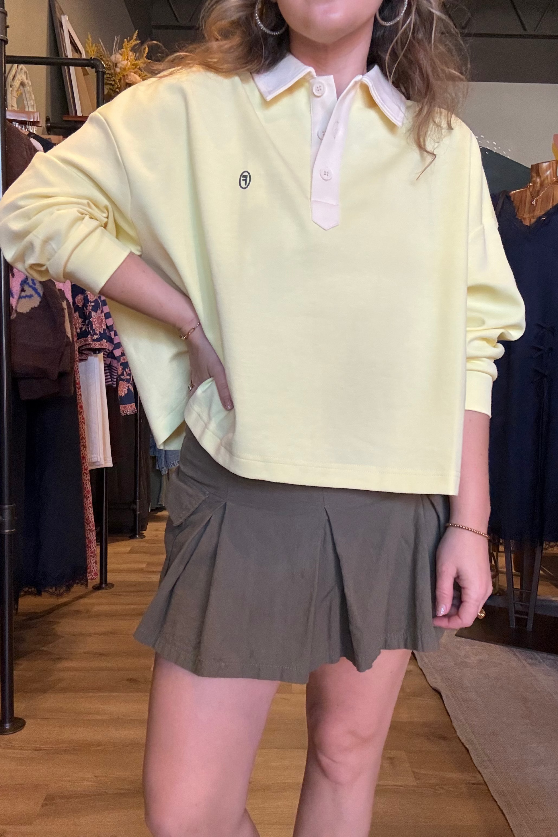 A person wearing a pale yellow long-sleeve polo shirt with a white collar and a pleated olive green mini skirt, standing inside a clothing store.