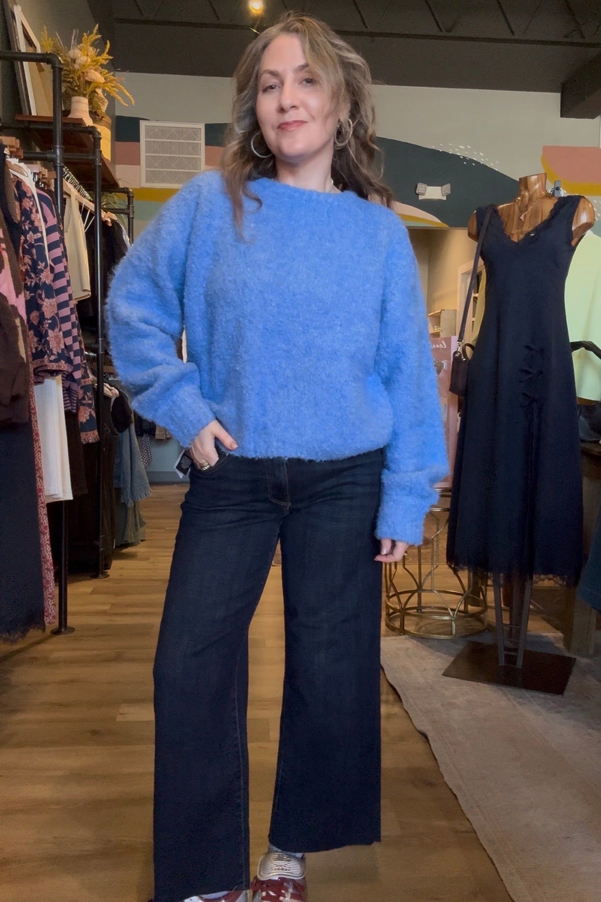 A woman stands in a clothing boutique wearing a fuzzy light blue knit sweater, dark wide-leg jeans, and white sneakers with red accents.