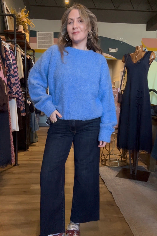 A woman stands in a clothing boutique wearing a fuzzy light blue knit sweater, dark wide-leg jeans, and white sneakers with red accents.