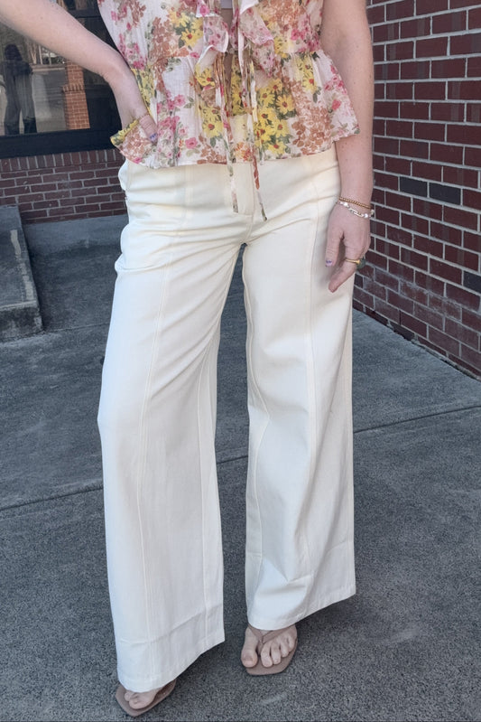 Person wearing a floral blouse and white pants standing against a brick wall.