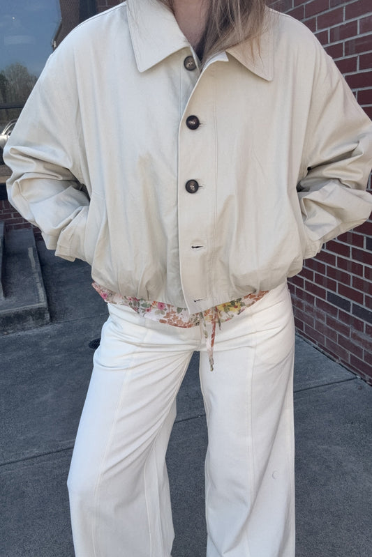 Person wearing a beige jacket and white pants with a floral shirt underneath, standing on a sidewalk.