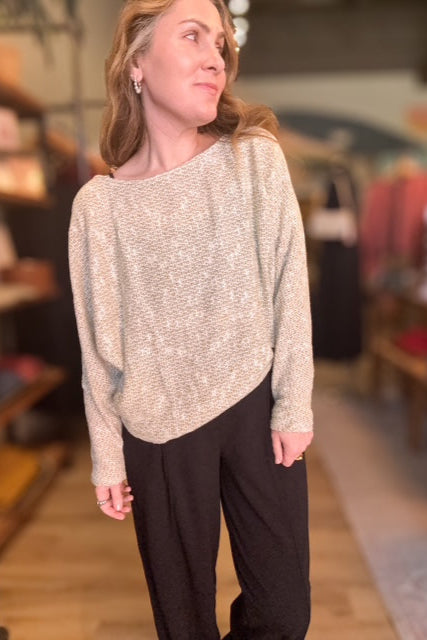 Woman wearing a beige sweater and black pants in an indoor setting with shelves and decor.