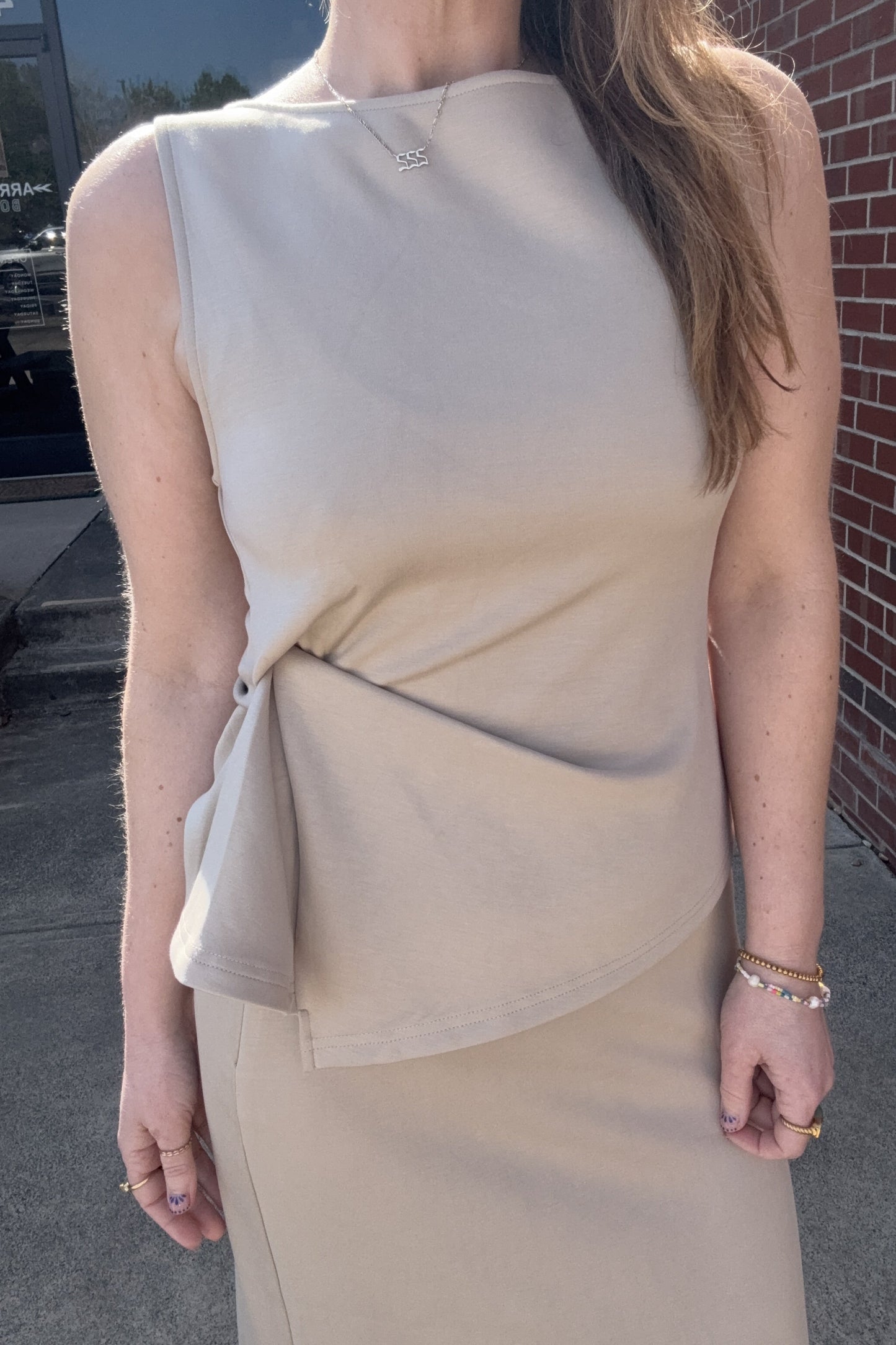 Woman wearing a beige sleeveless top with a knot detail, standing outdoors near a brick wall.