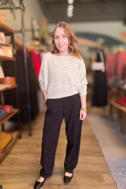 Woman standing in a clothing store wearing a light sweater and dark pants.