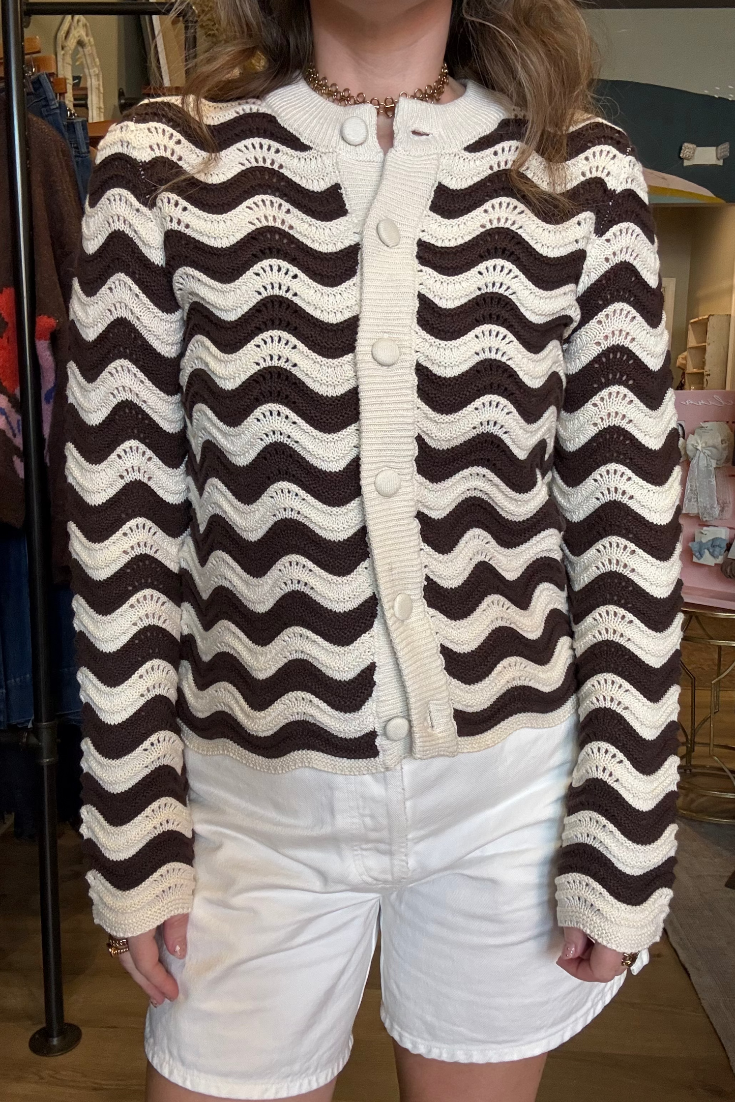 A person wearing a cropped knit cardigan with a brown and cream wavy stripe pattern, paired with white shorts, photographed in a boutique setting.